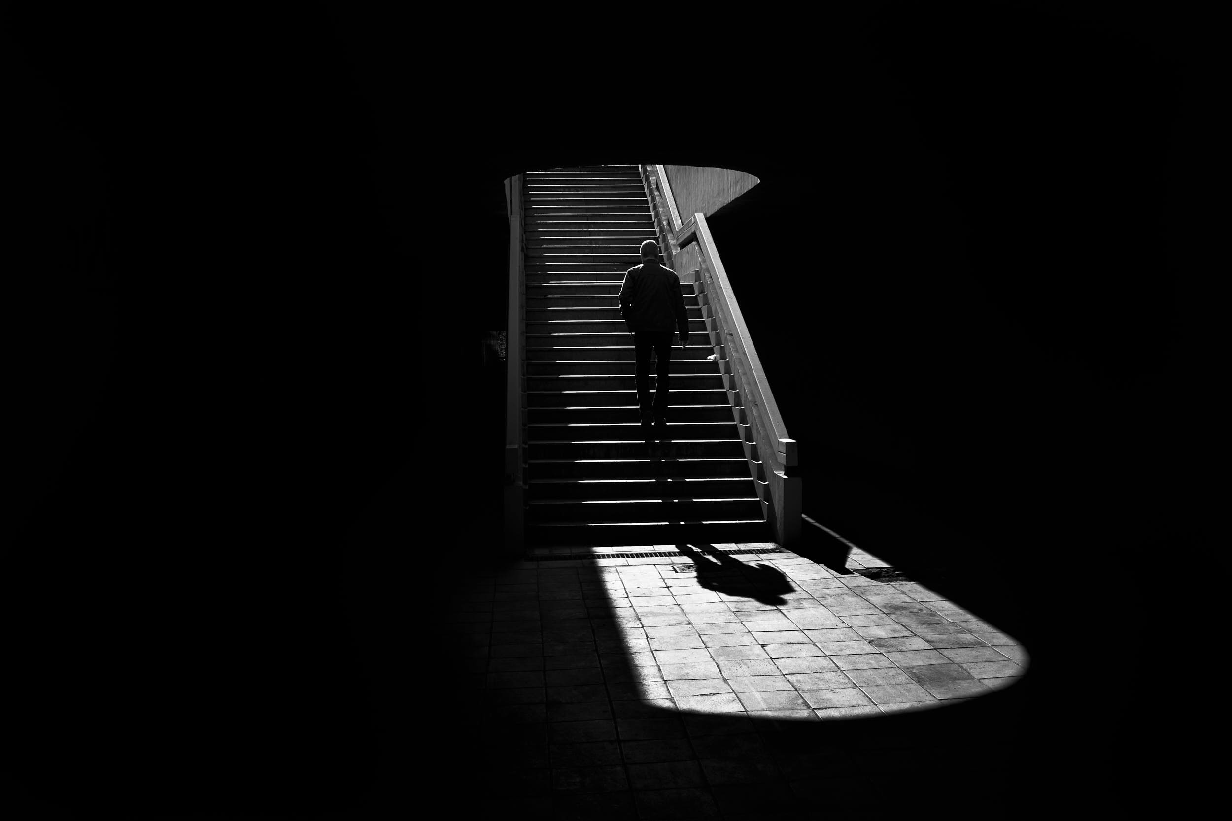 Black and white street photography: man walking up a staircase in an urban scene with deep shadows and strong contrast.