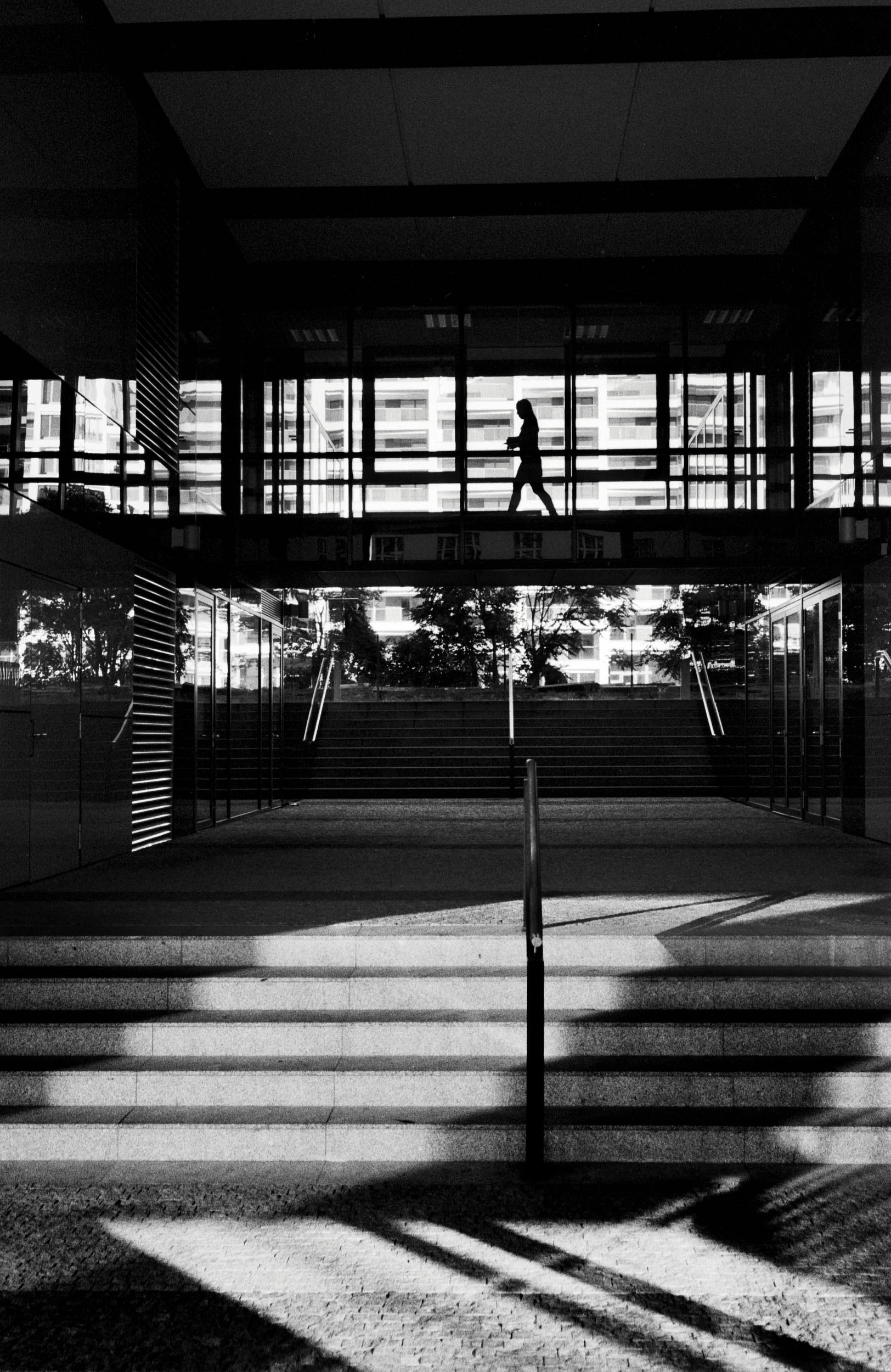 Classic black and white street photography: silhouette of a woman walking through a glass corridor in an office building.