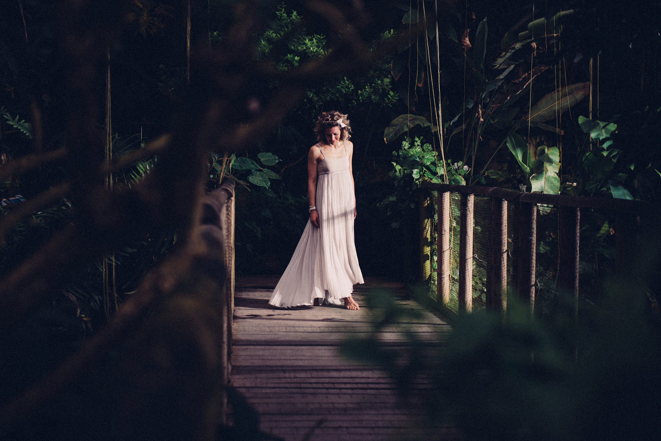 Woman in minimalistic dress walking across a wooden bridge in a jungle-like scene.