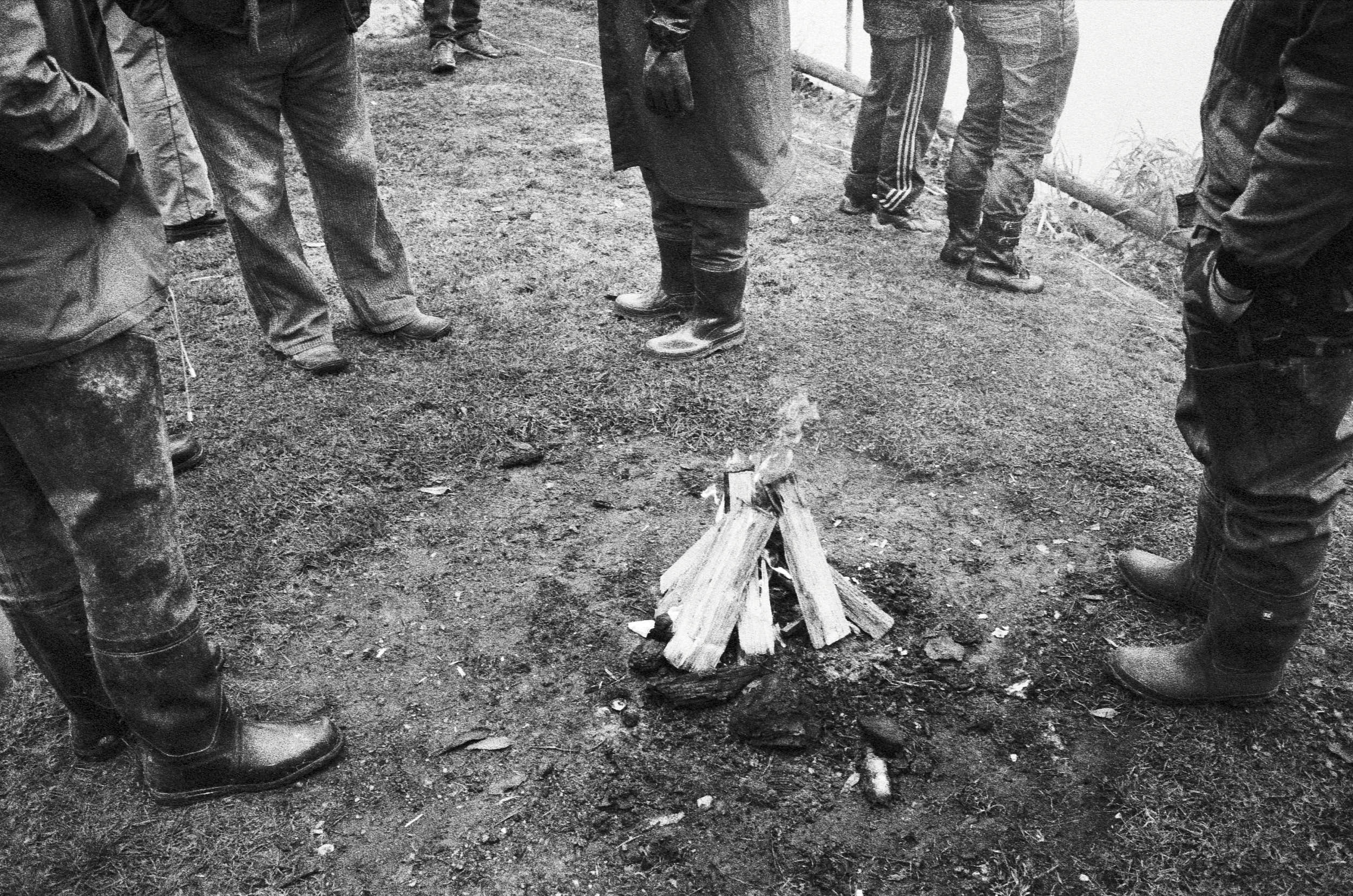 Group of people in wellington boots gathering around a campfire in a black and white documentary photograph.