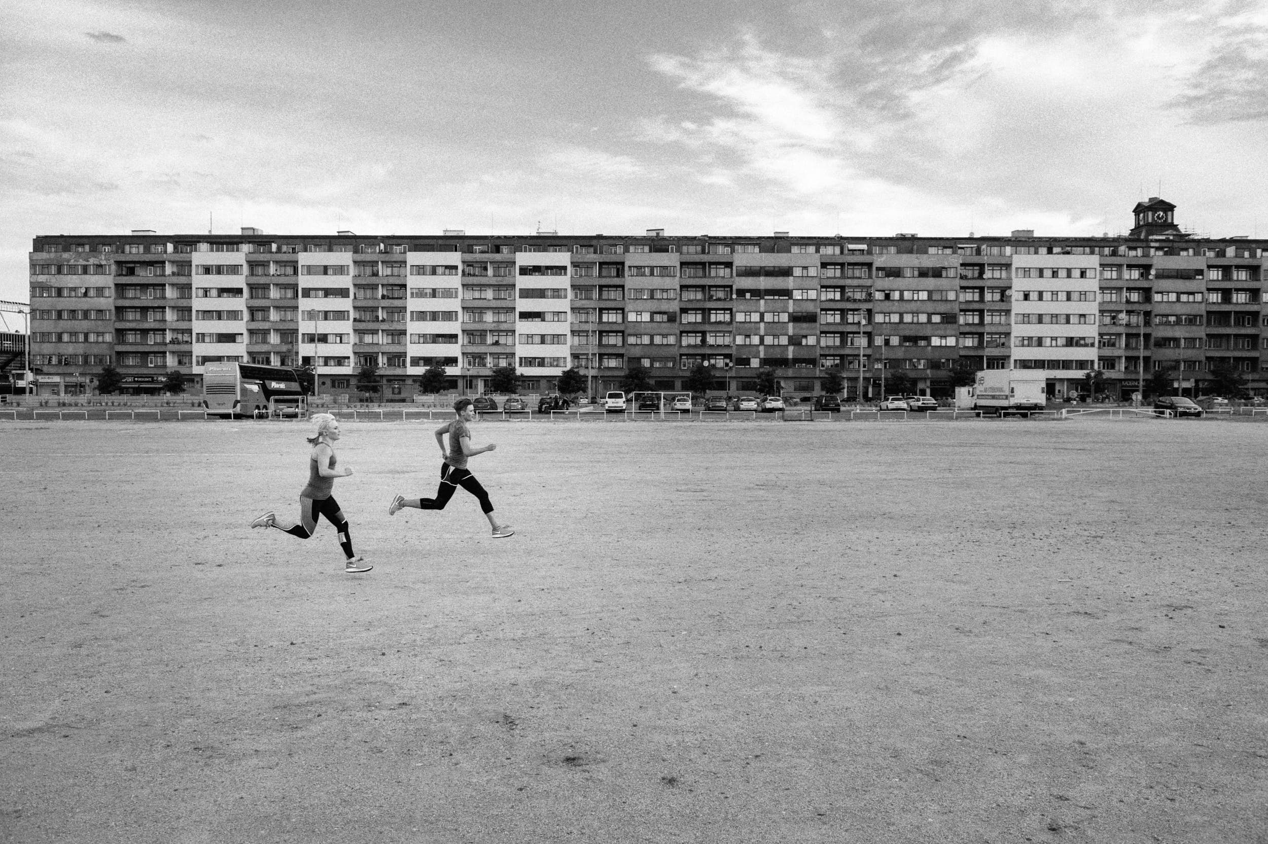 Black and white sport photography: two girls running across a sand plain with big housing estate in the background.