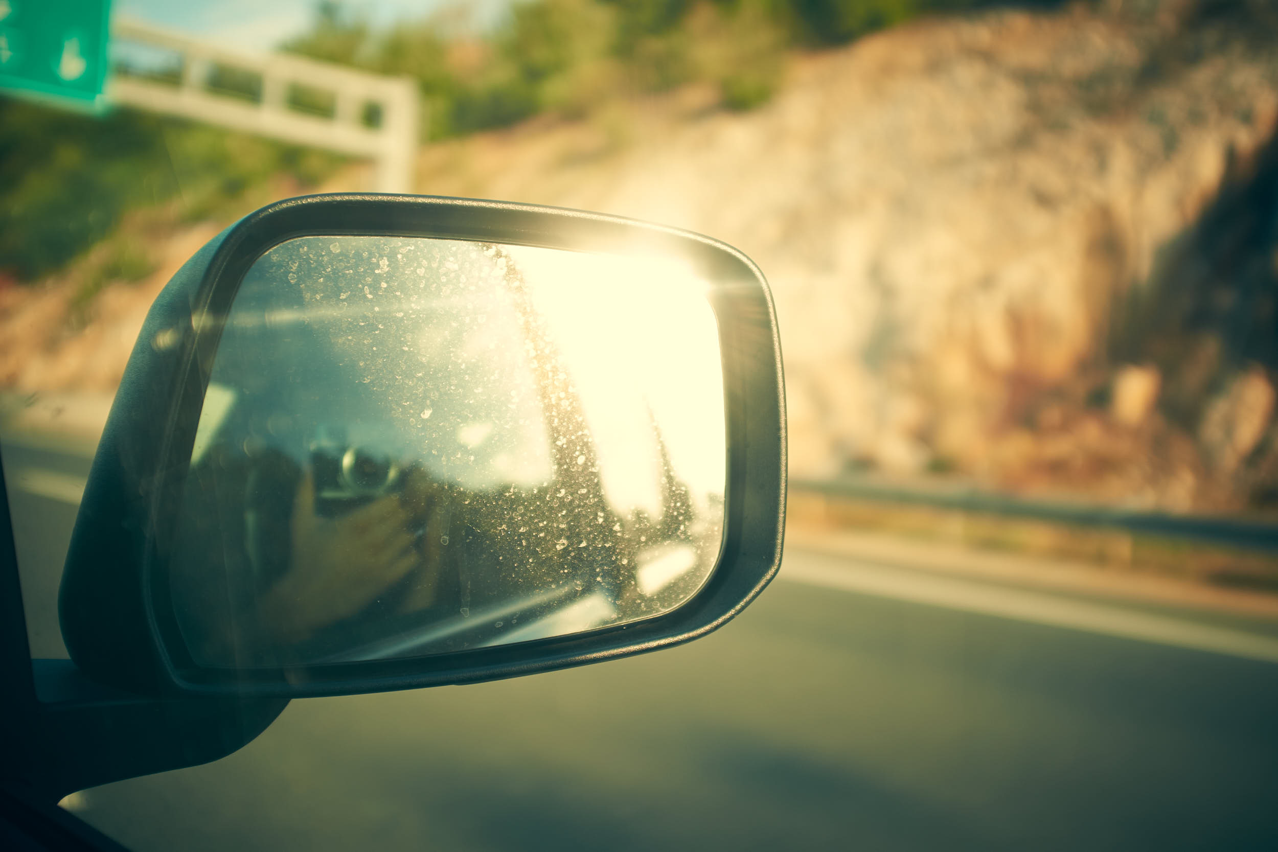 Travel photography on the road. Rearview mirror reflecting sun flare with blurry highway in the background.
